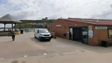 Google Public toilets on Pier Road in Whitby. A low-level brick building with a glass door and a white sign reading toilets in green letters