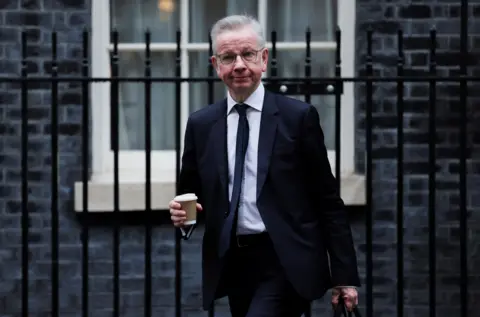 Michael Gove is seen walking along Downing Street. He is holding a takeaway coffee cup and is wearing a dark suit, white shirt and dark blue tie