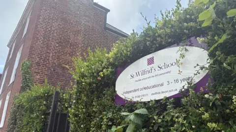 A purple and white sign for St Wilfrid's School set in a hedge at the entrance to the school with a red brick building in the background.