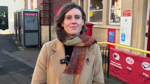 Jessica Toale stands outside the Westbourne Post Office, she it wearing a camel woolen smart coat, with a woolen scarf wrapped around her neck and over one shoulder in autumnal gingham colours like brown, red and orange and a black top underneath. She has short dark brown wavy hair in a side parting and smiles looking just off camera.