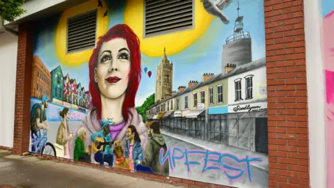 Upfest This mural has a woman with red hair standing with her head held high looking into the distance. Surrounding her are colourful buildings and shops. Underneath the woman is a group of people standing together. One person has a dog, another has a bike and one person is playing the guitar. 