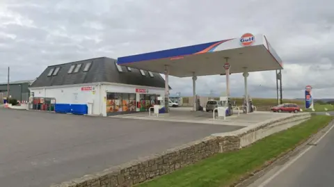 A Spar shop contained within a Gulf petrol filling station in Cornwall. A silver car is parked at one of the petrol pumps and a red car is driving onto the forecourt.