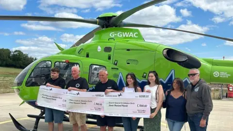 Andrew David The image shows a group of people standing in front of a Great Western Air Ambulance helicopter. The group are holding three large cheques, each for £7,000. 