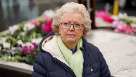 An older lady with a blonde perm, wearing a navy coat, green scarf and glasses looking to the left of the photo. 