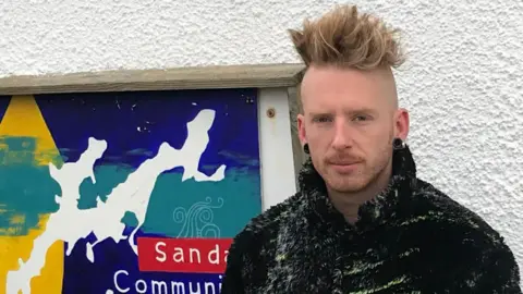 David Warner A man with a blonde quiff and black plug earrings stands next to a "Sanday Community Craft Club" sign. He is wearing a black and green fleece.