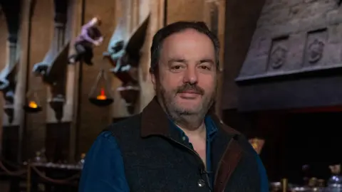 All characters and elements © & ™ Warner Bros. Entertainment Inc. Pierre Bohanna stands facing the camera in the Great Hall set at Leavesden Studios. He is shown from the chest up, wearing a blue shirt and a dark sleeveless vest. Behind him are the tall stone walls of the Great Hall, with carved details, sculpted figures, and wall-mounted torches glowing with warm light above long wooden tables and film props.