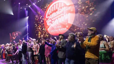 John Millard Photography A group of people on stage singing. They are standing in front of a red Sunday for Sammy sign which his lit up. There is a man at the front in a red and white Sunderland strip. Tim Healy is being shown on the screen to the left of the stage.