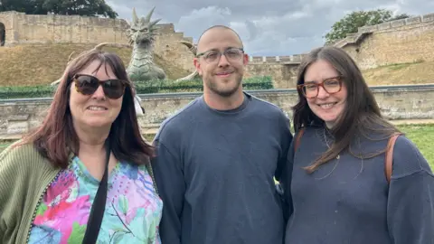 Eleanor Maslin/BBC Three people smiling at the camera with a large sculpture of a grey dragon in front of castle walls behind them. On the left, a woman with shoulder-length dark red hair is wearing large sunglasses and a colourful top. In the middle is a bald man with grey clear glasses and a navy jumper. On the right is a woman with long brown hair and thick brown clear square glasses, also wearing a navy jumper.
