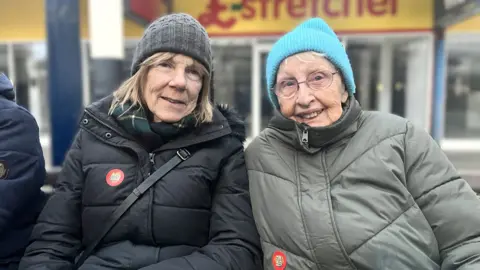 Maddy Jennings/BBC Heather English and Margaret Goodyear sat on a bench. Both are wearing padded coats and woolly hats. Heather has shoulder length light brown hair and Margaret has round rimless glasses.