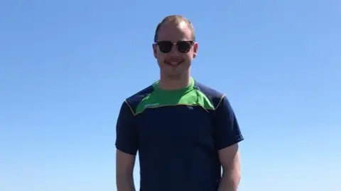 Sean Morgan Craig Ballentine smiling at the camera on a sunny day.  He has short, fair hair and a moustache.  He is wearing sunglasses and a navy and green T-shirt.