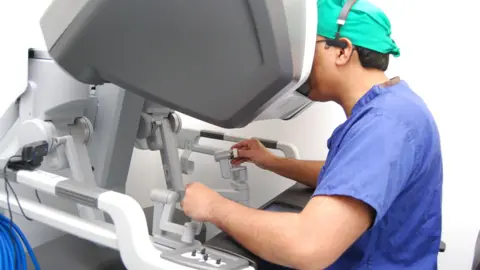 UHN Jamil Ahmed sits on a seat operating a robot. He is wearing scrubs and has a headset on. 