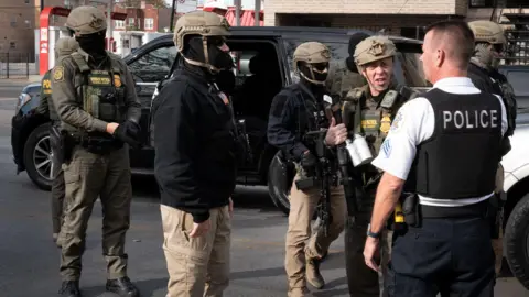 Getty Images A number of hooded and helmeted ICE agents wearing camo and green gear are stood near a large black car talking to a police officer.
