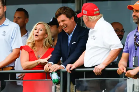 Getty Images Marjorie Taylor Greene in a red dress brushing against a laughing Tucker Carlson in a suit, listening to Donald Trump who has his backed turned to the camera and is speaking to the other two. 