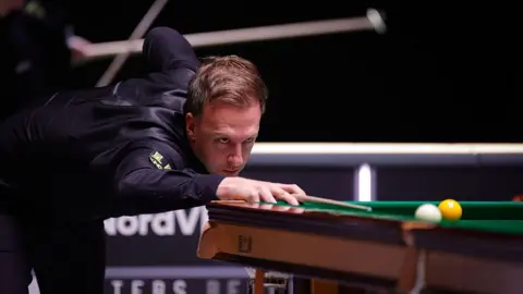 Judd Trump lines up a shot. He has brown hair, brushed to his right, and is wearing a black shirt and waistcoat.