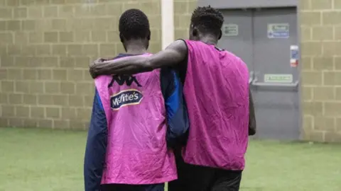 Lens Lab Project Two football players are stood with their backs to the camera. The slightly taller man has his arm around the other man's shoulder. Both men are wearing pink jerseys and black shorts.