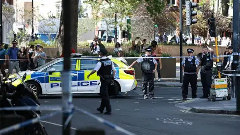 A group of police gather around the scene with is covered in police tape alongside a police vehicle.