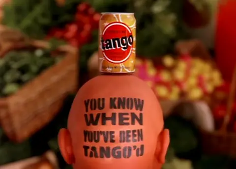 Screen grab showing a back-of-the-head shot of the "Orange Man" character from the iconic 1992 Tango Orange advertisement. A vintage orange Tango can rests on top of his bald, orange-painted head, which is stamped with the classic brand slogan: "You Know When You've Been Tango'd"