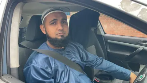 BBC/Seb Cheer A man with a dark beard and blue clothing sits in a car, looking down the camera lens through an open driver's window.