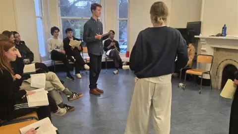 A group of students taking part in a class. They are sat in a circle of chairs with scripts in their laps. Two people are standing in the middle facing each other.