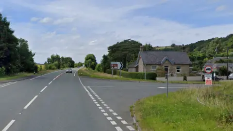 Google A single carriage road with a car approaching and a junction in the foreground with a sign to Embleton. There is a house on the corner and some more houses to its right.