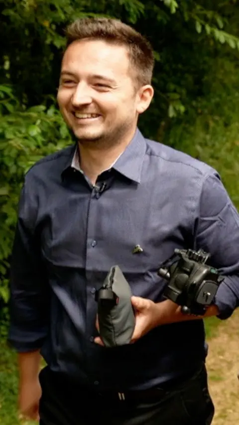 A man is wearing a blue shirt and is holding a camera while walking in wildlife.