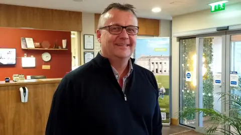 Terry is smiling into the camera. He's standing in a reception area. He wears glasses.