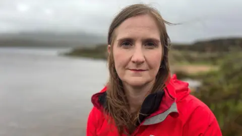 A woman with long brown hair and a red rain jacket. She is standing in front of a loch.