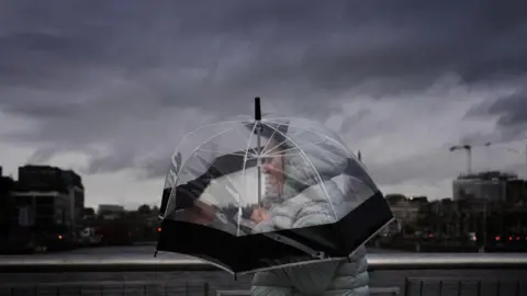 PA Media Dark storm clouds over Dublin. Someone wearing a winter coat with their hood up clutches their umbrella.