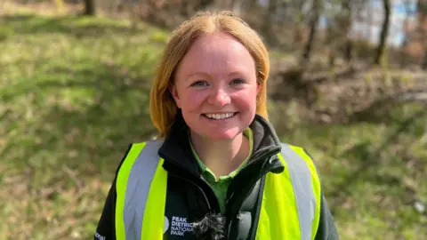 Woman with red hair and high vis jacket smiling infront of grassy verge 