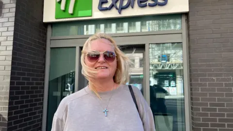 Jonny Humphries/BBC Amanda Thompson, who has shoulder length blonde hair and is wearing a ligh grey jumper with a crucifix necklace around her neck, smiles at the camera outside a Holiday Inn express entrance. Her wind is being rustled by the wind. 