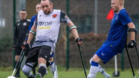 Courtesy of Cambridge City FC Football players with crutches are on a pitch during a match.