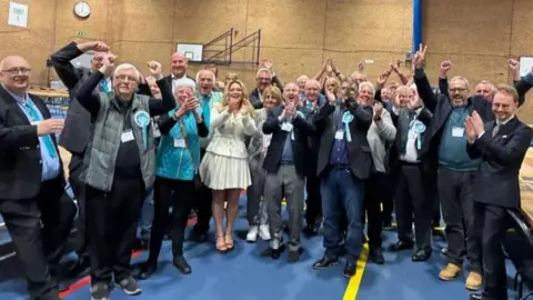 LDRS A group of men and women wearing the light blue rosettes of the Reform Party are standing in a group and cheering and clapping 