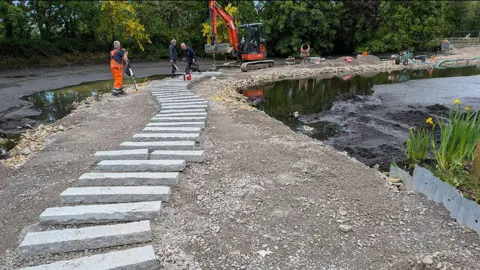 WWT Martin Mere Wetland Centre Workmen in orange overalls are working on laying stepping stones in a lake at a nature reserve. A digger is seen in the background. 