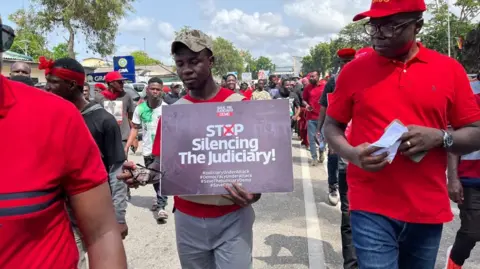 THOMAS NAADI / BBC Protesters march together, one of them holds a banner that reads: 'STOP silencing the judiciary'.