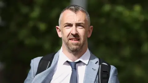 PA Media Daithí McKay has salt and pepper coloured hair and a beard and is wearing a light grey suit, white shirt and dark coloured tie