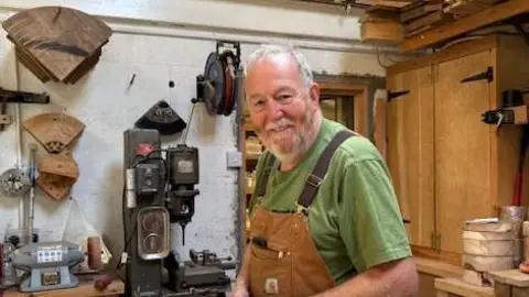 DEBRA TRACEY Graham Hall, a man with a grey beard looks at the camera, he is stood in his woodwork studio. 