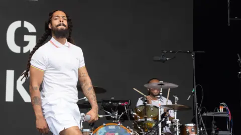 Getty Images Bobby Vylan, a man with long brown hair and dressed in a white polo t-shirt and shorts, performs on stage at Glastonbury.