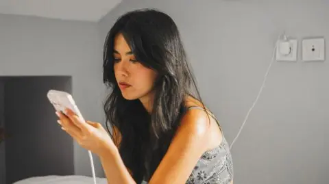 AFP via Getty Images A woman uses a smartphone while charging it in a bedroom. She is sitting on a bed and the phone cable is plugged into a wall socket behind her. She has long black hair and is wearing a top with straps, and she is sitting looking at the screen.