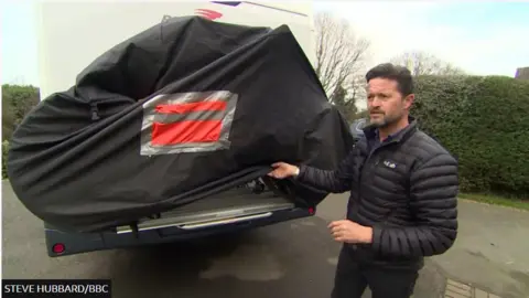 Steve Hubbard/BBC A man in a black puffer jacket standing next to his motorhome.