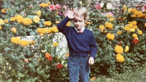 A grainy film photograph from the 1980s showing a young boy, perhaps aged about four, waving as he stands in a garden. He is wearing a white shirt, red tie, navy jumper and blue cords. Behind him are lots and lots of flowers, mostly yellow although there are red, orange and pink flowers too, and a Cotswold stone wall.