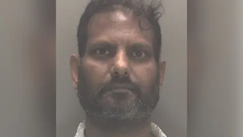 Police custody image of Nimalarajah Mathiyadaranam with black hair and beard wearing a white patterned shirt. 