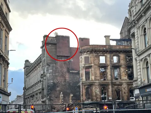 The fire-ravaged shell of a building on the corner of a street in the centre of Glasgow. There is a chimney at the top of the building, which was previously attached to the building which has now collapsed. It appears to hang in mid-air and has a red circle around it.
