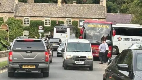 A traffic jam of cars, vans and a coaches on a road through a typical Cotswold village