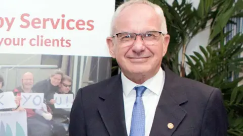 Dorset Community Foundation A smiling Tom Flood, wearing a navy blue suit, white shirt and pale blue tie. He has white hair and is wearing glasses.