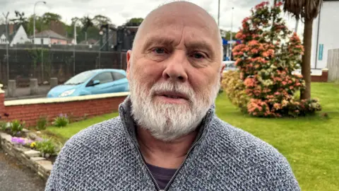 A man with a white beard. He has a grey half zip