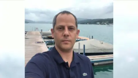 Eddy Eliaz is pictured looking straight into the camera. He is wearing a dark blue T-shirt and is standing on a jetty, which appears to lead out into a lake. There are speedboats moored next to the jetty and hills can be seen in the distance. Mr. Eliaz has dark eyes and dark, short hair.