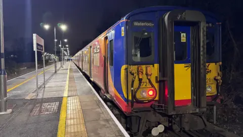 A red South Western Railway Class 455 train at a station platform, with a yellow and blue front.