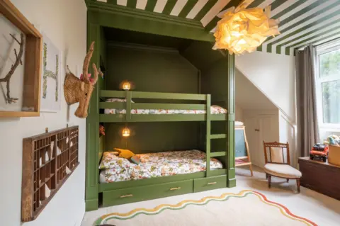 IWC Media A cosy children’s bedroom features a green built-in bunk bed with patterned bedding and warm wall lights tucked into the frame.
Playful decor, including a woven animal head, framed artwork, toys, and a soft rug, creates a bright and inviting space near a large window.