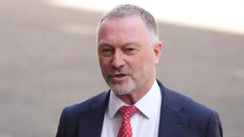 PA Media Steve Reed, a man with short grey hair and beard in a blue suit with a white shirt and red tie