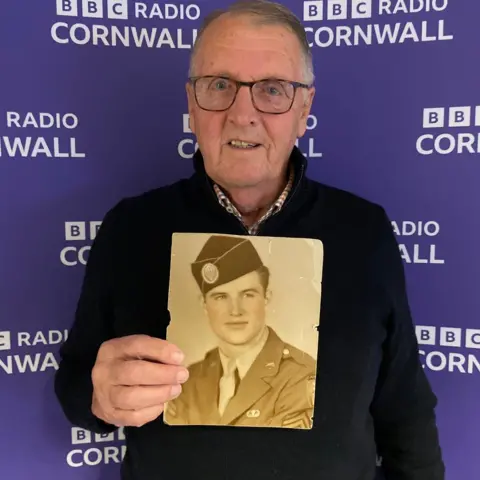 Tim Lobb is wearing glasses, a black jumper and collared shirt and is holding an old photograph of his father. He is standing in front of a BBC Radio Cornwall screen.